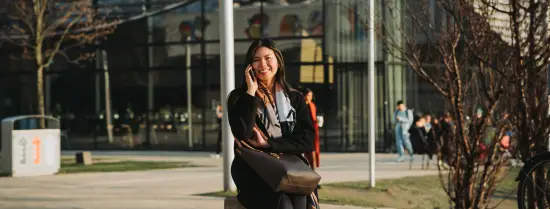Een jonge vrouw zit buiten op een stenen krukje te telefoneren en lacht naar de camera