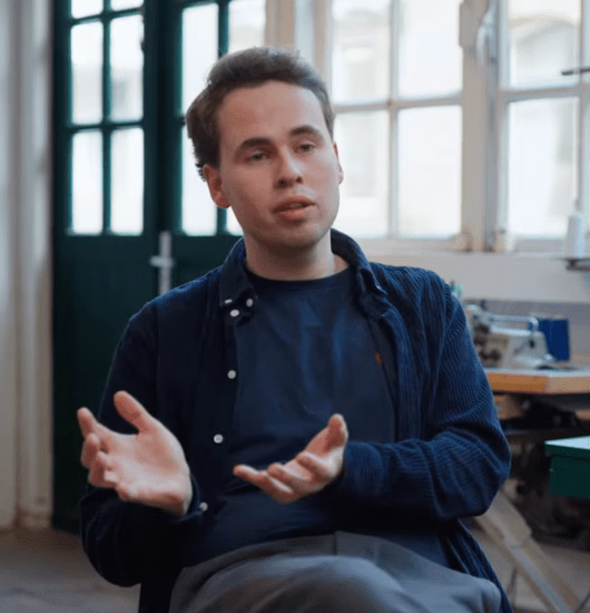 Young man in blue shirt sits indoors, gesturing with hands by a window.