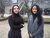 Two women in winter clothes standing outdoors by bare trees and a glass building.
