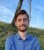 Man in blue shirt stands on grassy hill under blue sky with tree behind.