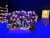 Theater cast in black gathered center stage under blue lights, props shelf left, yellow sign right.
