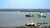 Green container ship crossing a wide river with tugboats near the riverbank under cloudy sky