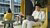 Photo of man in light suit using laptop in yellow lounge chair, woman reading behind him