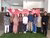 Nine colleagues standing on indoor turf, smiling in front of a red event banner.