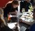 Photograph of a woman writing at a café table with documents, wine glasses and colleagues.