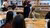 Students sitting and eating in a round table while listening to a woman talk