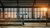 A train platform at Berlin Friedrichstraße station with a modern glass roof through which the sun shines, and some people in the background.