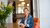 White woman with short hair sitting at a dining table with orange chairs. Flowers on the table.