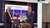 Two executives in suits exchange signed documents in front of a digital display screen.