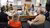 Eight colleagues seated around an oval table in a sunlit office talking over laptops and documents.