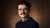 Man with dark hair, glasses and mustache wearing navy sweater against brown background