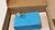 Open cardboard box holding a blue gift box tied with string and small silver padlock