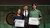 Woman and man holding certificates beside suited man on stage with green backdrop