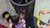 Two children searching inside a trash bin on concrete while an adult hand offers a yellow cloth.