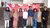 Nine colleagues standing on indoor turf, smiling in front of a red event banner.