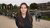 Smiling woman with long brown hair in dark hoodie on walkway by bikes and flags