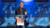 Man in dark jacket speaking and gesturing at transparent podium on blue stage