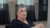Oudere vrouw met kort grijs haar en zwart shirt, zittend in interviewruimte met gordijnen