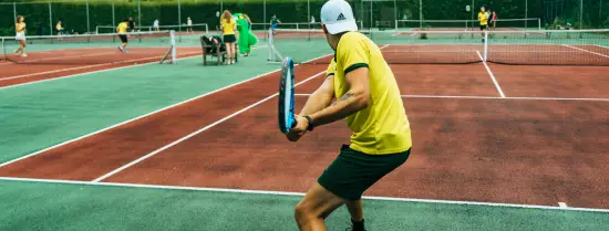 Persoon van achteren gezien in geel T-shirt met tennisracket op tennisbaan.