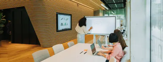 Two people at a meeting table, watching someone draw on a smart screen.