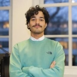 Man in mint green sweatshirt standing arms crossed by a window.